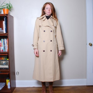 Puede incluir: Una gabardina larga de color beige con cierre de doble botonadura y botones marrones. La gabardina tiene un cinturón y se lleva con botas marrones.