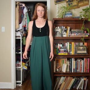Könnte beinhalten: Eine Frau trägt ein schwarzes und grünes Maxikleid mit einem Knopfbodice. Sie steht vor einem Bücherregal voller Bücher.