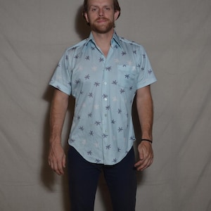 May include: A light blue short-sleeved shirt patterned with white and grey star shapes. The shirt has a button-down front and a chest pocket. The person is wearing navy blue trousers.