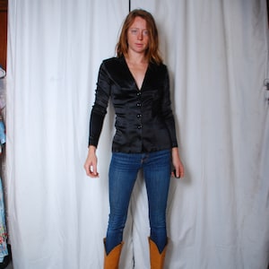 May include: A woman wearing a black satin button-up shirt with long sleeves and blue jeans. She is wearing brown cowboy boots.