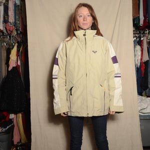 May include: A tan and white zip-up jacket with purple sleeve detailing. The jacket features a high collar and a small logo. The person is wearing dark blue jeans and black boots. Clothing.