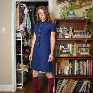 Puede incluir: Una mujer con un vestido azul de cuello con costuras blancas y botas de color burdeos hasta la rodilla. Está de pie frente a una estantería llena de libros y un armario.