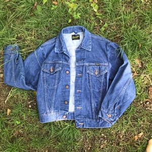 Puede incluir: Una chaqueta vaquera azul con una etiqueta de Wrangler en el bolsillo. La chaqueta tiene un dise&ntilde;o cl&aacute;sico con cierre de botones en la parte delantera y dos bolsillos con solapa.