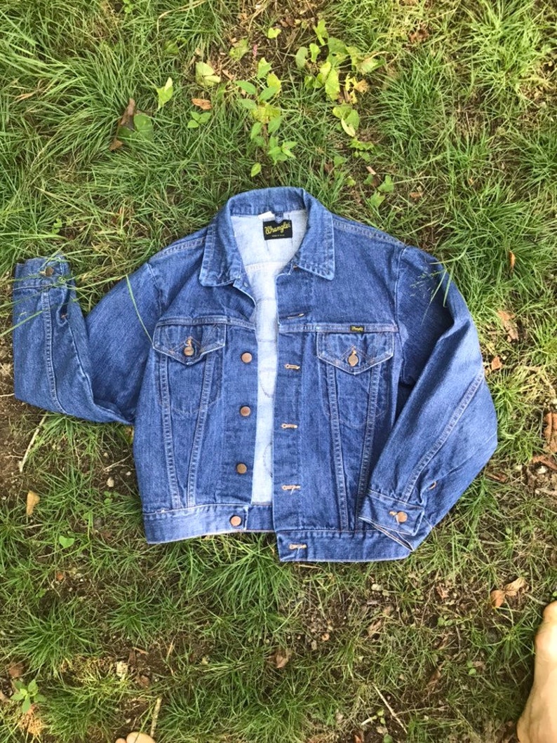 Puede incluir: Una chaqueta vaquera azul con una etiqueta de Wrangler en el bolsillo. La chaqueta tiene un dise&ntilde;o cl&aacute;sico con cierre de botones en la parte delantera y dos bolsillos con solapa.