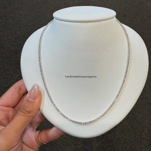 May include: A silver-toned tennis necklace featuring a continuous row of small, clear, round-cut stones set in a metallic chain. The necklace is displayed on a white, rounded neck form. The text handmadetreasuregems is printed in the center.