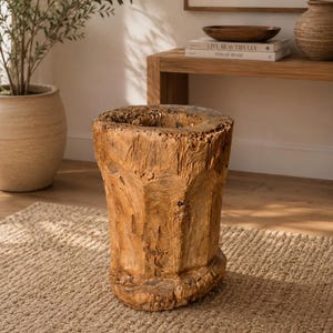 Op de afbeelding: Een cilindrische houten kruk met een ruwe, natuurlijke textuur en een lichtbruine afwerking. Het object heeft een uitgeholde bovenkant, zichtbare houtnerven en een stevige, dikke basis met een onregelmatige, organische vorm en verweerde details.