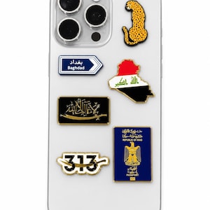 May include: A clear smartphone case decorated with several enamel pins including a cheetah, a Baghdad road sign, the map of Iraq with its flag, a black rectangular pin with gold Arabic calligraphy, the number 313, and a blue Republic of Iraq passport.