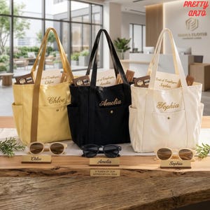May include: Three canvas tote bags in yellow, black, and white, each featuring a front pocket with a name embroidered in script: Chloe, Amelia, and Sophia. Each bag includes a pair of sunglasses and a small nameplate placed on the wooden surface.