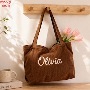 May include: A brown corduroy tote bag featuring the name Olivia in white script lettering on the front. The bag has two shoulder straps and contains three light pink tulip flowers. The fabric has a vertical ribbed texture throughout the exterior.