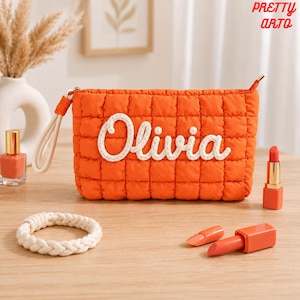 Op de afbeelding: Een rechthoekig oranje gewatteerd tasje met de naam Olivia in witte touwachtige letters op de voorzijde. Het tasje is voorzien van een bijpassend oranje polsbandje en een ritssluiting. Het ontwerp is eenvoudig, functioneel en helder van kleur.