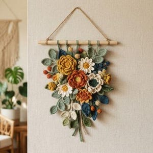 May include: A wall hanging featuring a crochet floral arrangement in shades of mustard yellow, burnt orange, cream, and sage green. The flowers and leaves are attached to a horizontal wooden dowel suspended by a thin, light brown twine cord.