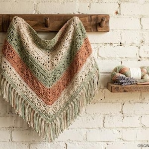 May include: A triangular crochet shawl featuring horizontal stripes in sage green, terracotta, and cream with an intricate lace pattern and long fringe along the bottom edge. Two balls of yarn and a crochet hook sit on a small wooden shelf nearby.