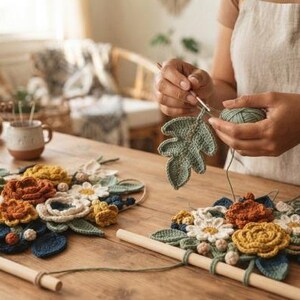 May include: Two wall hangings featuring crocheted flowers in shades of rust, mustard yellow, white, and dark blue, attached to wooden dowels. A crocheted green leaf is being worked on with a hook, with yarn spools visible on the table.