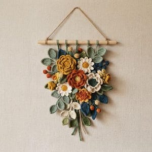 May include: A wall hanging featuring a collection of crocheted flowers and leaves in shades of mustard yellow, burnt orange, sage green, and navy blue. The arrangement is suspended from a horizontal wooden dowel with a thin twine cord.