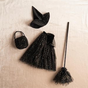 Puede incluir: Un conjunto de disfraz de bruja negro que incluye un sombrero puntiagudo, una falda, un bolso pequeño y una escoba. Cada pieza está confeccionada en tela negra con un patrón de estrellas doradas. La falda es de tul en capas y la escoba tiene un mango largo.