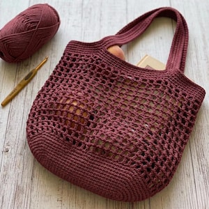 May include: A dark mauve crochet tote bag featuring a mesh body with a solid base and two integrated handles. Beside the bag sits a matching ball of yarn and a gold-toned crochet hook, showcasing the construction of this handmade textile accessory.
