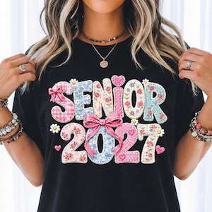 Könnte beinhalten: Ein schwarzes T-Shirt mit dem Schriftzug SENIOR 2027 in großen Buchstaben und Zahlen. Die Zeichen sind mit verschiedenen Blumen-, Vichy- und Punktmustern in Rosa, Blau und Creme verziert und mit einer großen rosa Schleife akzentuiert.