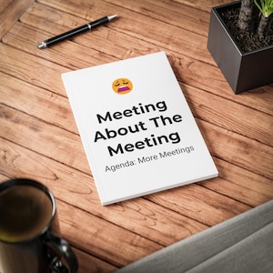 Puede incluir: Un cuaderno blanco con una cara emoji amarilla en la parte superior, con el texto negro Meeting About The Meeting y el subtítulo Agenda: More Meetings impreso en la portada, colocado sobre una superficie de madera con un bolígrafo y una taza de café.