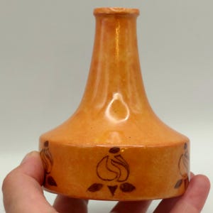 May include: A small, orange ceramic vase with a wide, flat base and a narrow neck. The surface features a mottled, textured finish in shades of light and dark orange, decorated with a repeating pattern of dark brown, hand-painted floral motifs.