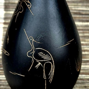 Puede incluir: Un jarrón negro mate en forma de lágrima con delicados dibujos de líneas grabadas en color dorado que representan aves de cuello largo. El jarrón tiene un cuello estrecho con un borde dorado fino en la parte superior y un cuerpo redondeado.