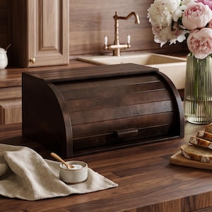 Puede incluir: Una panera de madera de color marrón oscuro con tapa enrollable sobre una encimera de madera. Al lado hay un pequeño cuenco de cerámica con una cuchara de madera, un paño de lino, un jarrón de cristal con peonías y pan en rodajas.