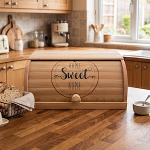 Könnte beinhalten: Ein hellfarbener Brotkasten aus Holz mit einem Rolldeckel, der ein kreisförmiges Design und den schwarzen Schriftzug "HOME Sweet HOME" auf der Vorderseite aufweist. Der Brotkasten hat einen kleinen runden Holzknopf zum Öffnen des Deckels.
