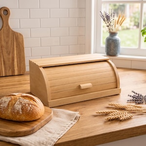 Könnte beinhalten: Ein hellfarbener Brotkasten aus Holz mit einem Rolldeckel und einem kleinen rechteckigen Griff. Der Brotkasten steht auf einer Holzoberfläche neben einem runden Laib Brot auf einem Schneidebrett, getrockneten Weizenähren und Lavendelzweigen.