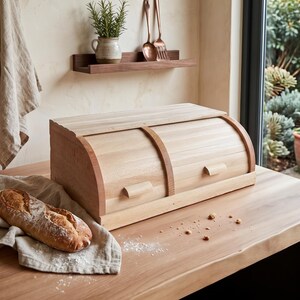 Puede incluir: Una panera de madera de color claro con un diseño de doble tapa enrollable y dos pequeñas asas. Una hogaza de pan crujiente descansa sobre un paño de lino beige cerca, con migas esparcidas sobre la superficie de madera de la encimera.