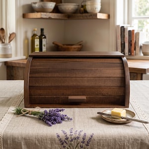 Puede incluir: Una panera de madera de color marrón oscuro con tapa enrollable y un pequeño tirador rectangular. La panera está colocada sobre una mesa junto a un pequeño ramo de lavanda morada y un plato de cerámica con un trozo de mantequilla.