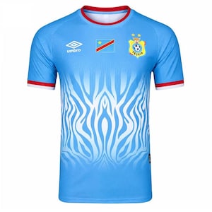 May include: A light blue short-sleeve jersey featuring a white zebra-like pattern on the front, a red crew neckline, and red sleeve cuffs. The shirt displays the Umbro logo, the flag of the Democratic Republic of the Congo, and the FECOFA emblem.