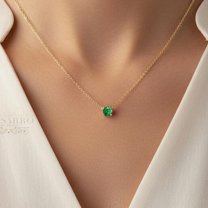 Könnte beinhalten: Eine zarte Goldkettenhalskette mit einem einzelnen, kleinen, rund geschliffenen grünen Edelsteinanhänger, der mittig auf dem Schlüsselbein sitzt. Die Kette wird zu einer schlichten weißen Bluse mit einfachem Ausschnitt getragen.