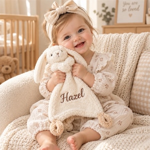 May include: A cream-colored plush bunny security blanket featuring a soft, textured fabric and long floppy ears. The blanket is embroidered with the name Hazel in dark brown script lettering and includes two small knotted loops at the bottom corners.