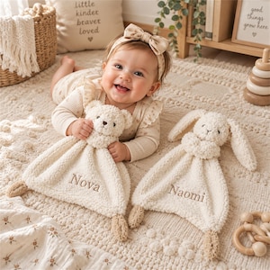 Può includere: Due doudou in peluche color crema con nomi ricamati. Uno ha una testa di orsetto ed è ricamato con il nome Nova, mentre l'altro ha una testa di coniglietto ed è ricamato con il nome Naomi. Entrambi sono realizzati in tessuto morbido e testurizzato.