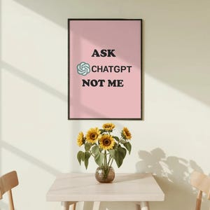 May include: A rectangular wall art print in a black frame featuring a light pink background with the black text ASK, the ChatGPT logo, and the text CHATGPT NOT ME. The print is displayed on a wall above a table with a glass vase of sunflowers.