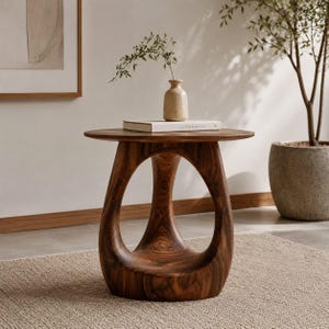 Può includere: Un tavolino rotondo in legno con venature marrone scuro e una base scultorea composta da tre gambe curve che si uniscono al centro. Un vaso in ceramica beige con un piccolo ramo di foglie verdi è appoggiato sopra un libro bianco intitolato The Kinfolk Home.