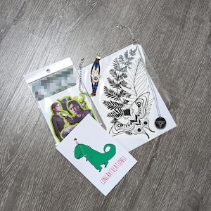 Puede incluir: Una colección de artículos que incluye una pulsera de cuero marrón con un dije de hamsa plateado, un collar plateado con un colgante circular, una pegatina con dos figuras, un tatuaje temporal de polilla y helecho, y una tarjeta con un dinosaurio verde y el texto CONGRATULATIONS.