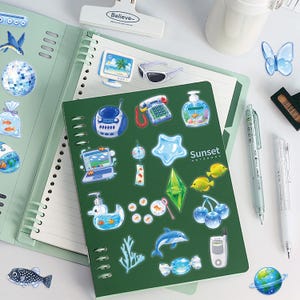 May include: A dark green spiral-bound notebook with the text Sunset Notebook on the cover, decorated with various blue and green stickers including a dolphin, fish, computer, telephone, and cherry icons, alongside a light green open notebook.