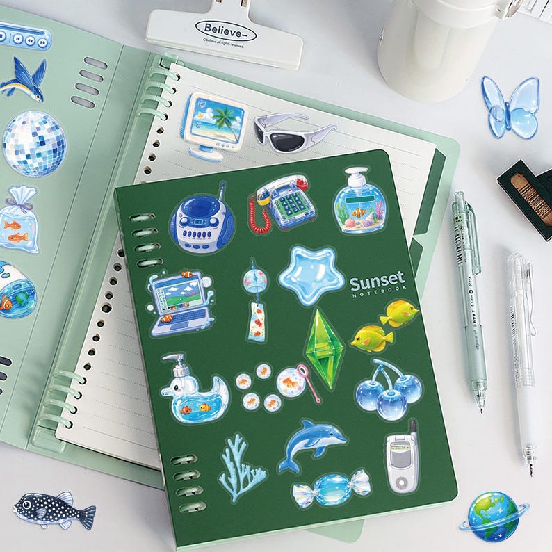 May include: A dark green spiral-bound notebook with the text Sunset Notebook on the cover, decorated with various blue and green stickers including a dolphin, fish, computer, telephone, and cherry icons, alongside a light green open notebook.
