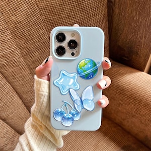 May include: A light blue smartphone case featuring a collection of raised, glossy stickers. The designs include a planet with rings, a five-pointed star, a butterfly, and a pair of cherries, all rendered in shades of blue and white.