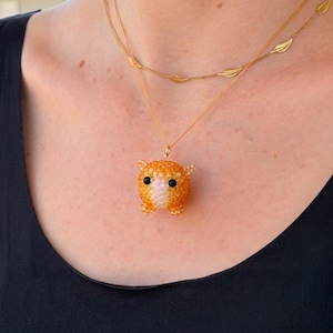 May include: A gold-toned necklace featuring a delicate chain with small leaf-shaped accents and a pendant crafted from tiny orange, white, and black beads shaped like a small, round cat with pointed ears and a light pink nose.