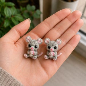 Pode incluir: Duas pequenas figuras de rato tridimensionais feitas de minúsculas contas cinzentas, brancas e cor-de-rosa. Cada rato apresenta orelhas redondas com centros cor-de-rosa, olhos de conta preta, um pequeno nariz rosa e bigodes brancos.