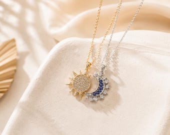 May include: A gold-toned sun pendant necklace featuring a circular center paved with clear stones and radiating rays, paired with a silver-toned crescent moon pendant necklace accented with alternating blue and clear stones on a delicate chain.