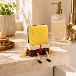 May include: A yellow rectangular sponge held by a brown plastic base with white trim, a red tie, and yellow legs with black shoes. The base features a small shelf design that supports the sponge upright, creating a character-like appearance.