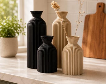 May include: A set of four fluted vases in black and beige. Two vases are tall and cylindrical, while two are shorter and wider. Each vase features a narrow neck and a vertical ribbed texture. One beige vase contains a delicate dried floral stem.