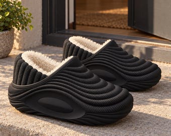 May include: A pair of black slip-on clogs featuring a thick, ribbed exterior texture and a smooth oval side panel. The interior is lined with a cream-colored, plush faux-fur material, providing a contrast to the dark, molded outer shell.