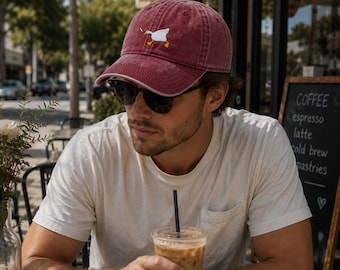 May include: A maroon baseball cap featuring an embroidered white goose with orange feet and beak on the front panel. The cap has a curved brim and a structured crown, constructed from a washed cotton fabric with visible stitching details.