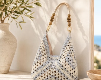 May include: A hobo-style handbag crafted from light blue and cream-colored crochet yarn in a geometric pattern. The bag features a single handle adorned with round wooden beads and light-colored circular rings connecting the handle to the body.