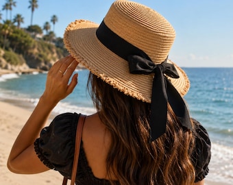May include: A tan straw hat with a flat top and a wide, frayed-edge brim. The hat is accented with a wide black ribbon band that wraps around the crown and ties into a bow with long, trailing ends hanging down the back of the hat.