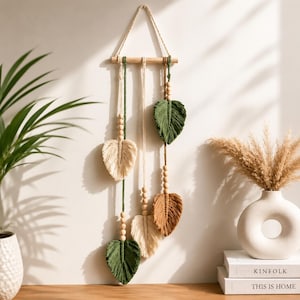 Op de afbeelding: Een macramé wandhanger met een horizontale houten stok en een ophangkoord. Drie verticale koorden in crème, groen en bruin hangen aan de stok, elk versierd met houten kralen en afgewerkt met een bladvormig kwastje van vezels.