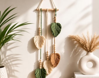 May include: A macrame wall hanging featuring a horizontal wooden dowel with a rope hanger. Three vertical cords in cream, green, and brown hang from the dowel, each decorated with wooden beads and finished with a leaf-shaped fiber tassel.
